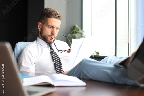 Handsome businessman sitting with legs on table and examing documents in office