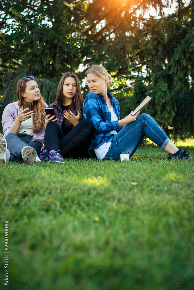 Three friends spend their free time sitting in the park on green lawn. Young pretty girls talking, smiling, looking photo at smartphone. Concept of female friendship. Vertical image. Three friends spend their free time sitting in the park on green lawn. Young pretty girls talking, smiling, looking photo at smartphone. Concept of female friendship. Vertical image.