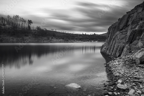 Riverscape on a cloudy early evening with water and rocks on foreground and a beach and trees on a hill in the background, Black and White Photo, Fronhas dam reservoir, Alva river, Fronhas, Portugal photo