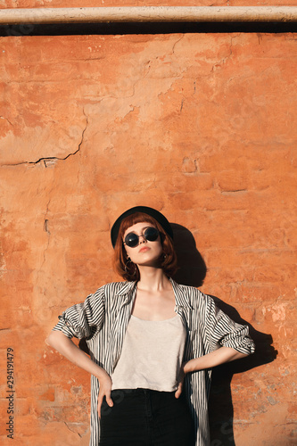 Young woman in striped shirt, hat and sunglasses posing near the orange wall