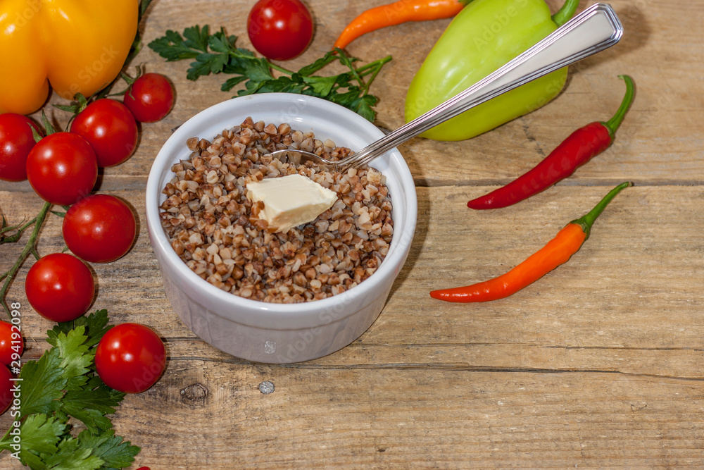 Cooked buckwheat porridge in a deep plate with butter on a wooden background with vegetables. Cooked buckwheat porridge in a deep plate with butter on a wooden background with vegetables.