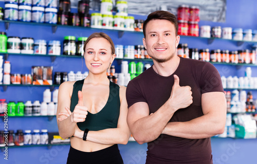 Young muscular people giving thumps up on background with shelves of sport nutrition products