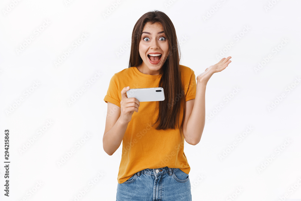Surprised, excited cute girl holding smartphone horizontally, raising one hand astonished, smiling happy and look camera telling she won game level, triumphing and rejoicing, white background Surprised, excited cute girl holding smartphone horizontally, raising one hand astonished, smiling happy and look camera telling she won game level, triumphing and rejoicing, white background