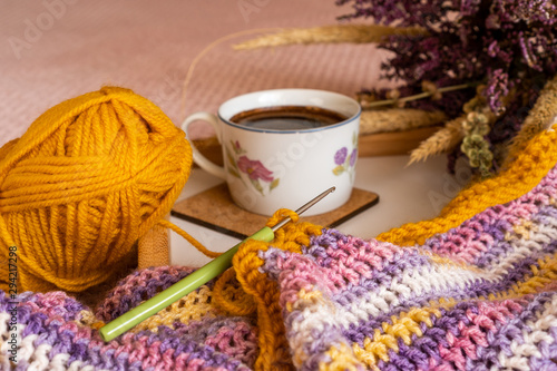Closeup of a colorful handmade infinity scarf in an autumn decor next to a cup of hot coffee and a bouquet of dried wildflowers. Double crochet pattern. Hand work in progress.