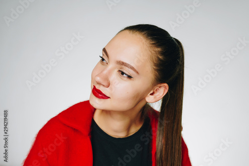 Amazing young girl with dark long hair tied in a ponytail, with red lipstick and in a red sweater with a black T-shirt on a white background, examines something aside with interest