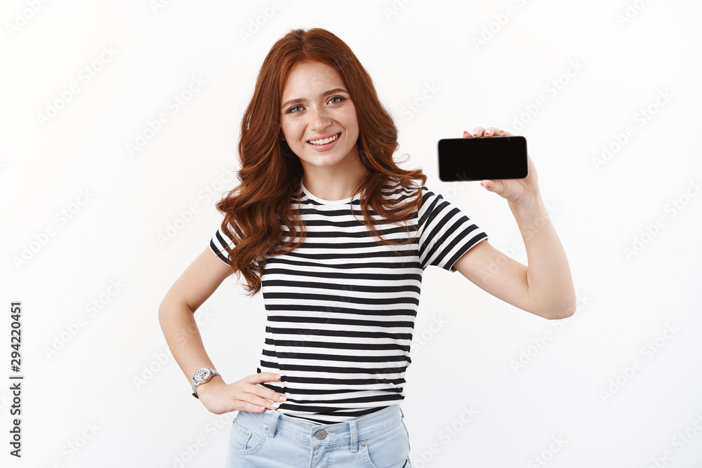 Cute redhead teenage female blogger accomplish goal, finish hard level, showing horizontally smartphone display, playing mobile phone game, smiling satisfied, standing white background Cute redhead teenage female blogger accomplish goal, finish hard level, showing horizontally smartphone display, playing mobile phone game, smiling satisfied, standing white background