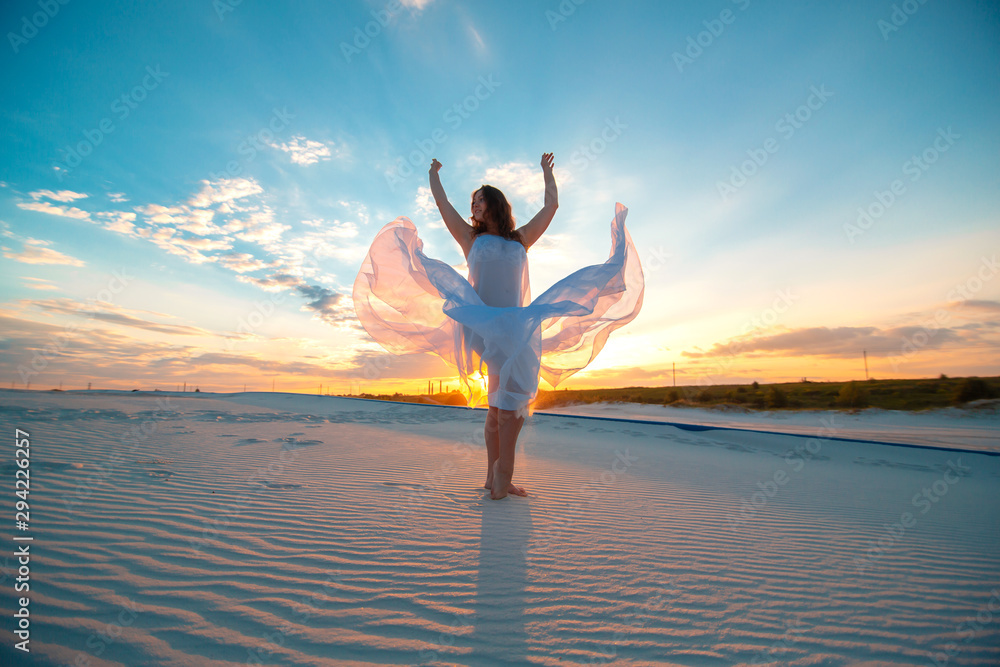 A girl in a fly white dress dances and poses in the sand desert at sunset.