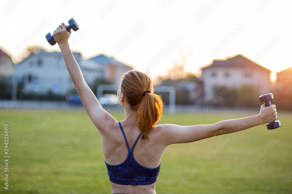 Portrait of athletic teenage girl in fitness wear exercising with blue dumbbells outdoors in park. Fit young woman working out outside.