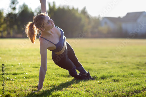 Young sportive woman in sports clothes training in field at sunrise. Girl standing in plank position on grass in a city park.