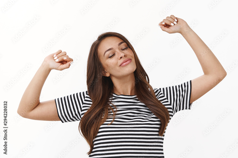 Obraz premium Waist-up shot carefree relaxed cute lazy girl stretching hands up, waking up energized, sleep well, close eyes and smiling delighted, enjoy weekends, standing in striped t-shirt white background