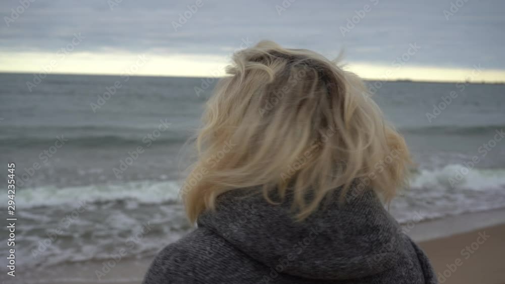 The girl turns and looks into the camera lens on the background of the cold sea. Blond hair in the wind. Hair and sea breeze. The girl turns and looks into the camera lens on the background of the cold sea. Blond hair in the wind. Hair and sea breeze.