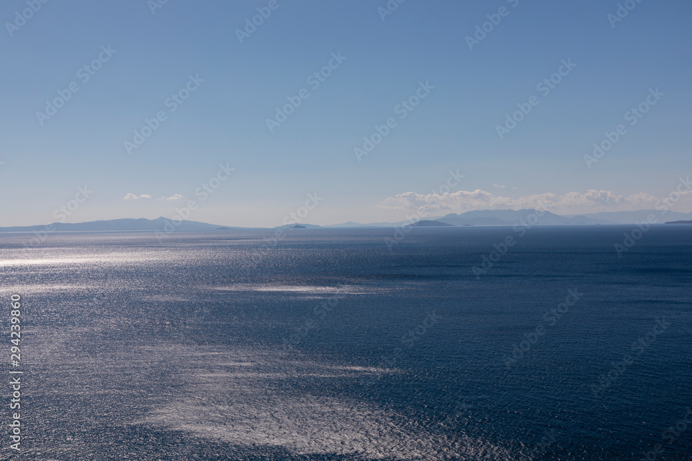 Bright blue ocean with mountains and clouds in the background and sun reflecting off the water.