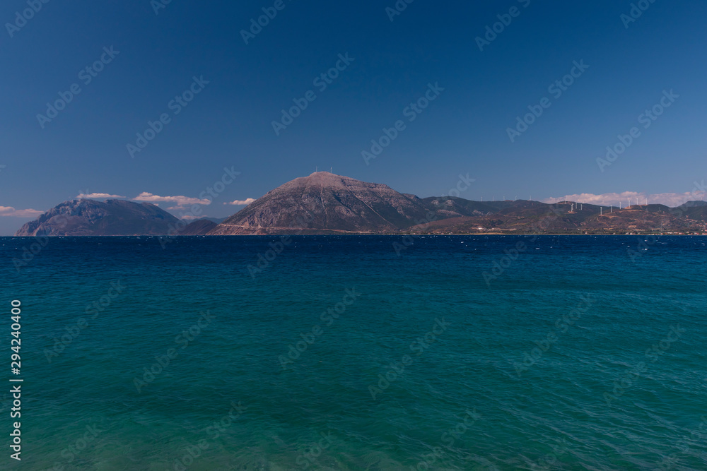 A mountain range in Greece with the ocean in the foreground and wind towers on the hill.