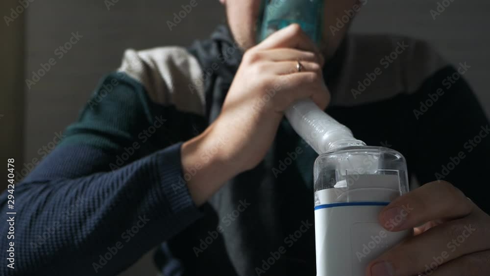 A sick man sits on a sofa in a sweater and with a scarf, uses an inhaler. Colds, illness, flu, treatment A sick man sits on a sofa in a sweater and with a scarf, uses an inhaler. Colds, illness, flu, treatment