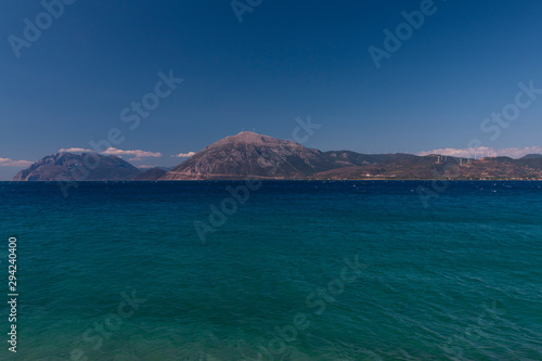 A mountain range in Greece with the ocean in the foreground and wind towers on the hill.