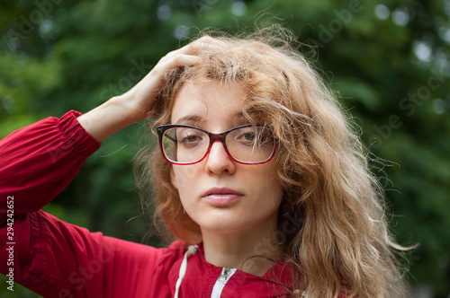 Young beautiful woman with blonde curly hair in dark red hipster jacket and eyeglasses posing outdoors in the urban park in the morning
