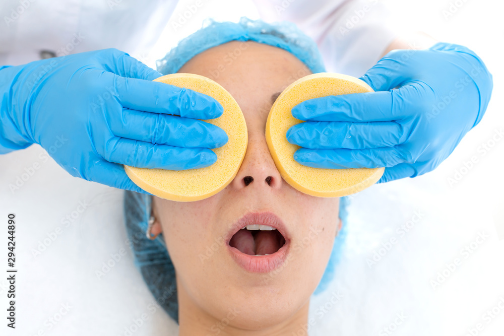 Naklejka premium A woman, a professional doctor, a beautician cleanses the patient's face with sponges before applying the mask for skin care