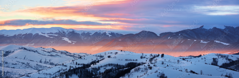 gorgeous panorama of mountainous countryside at sunrise in winter. snow covered rural area on rolling hills glow in morning sunlight. huge mountain ridge in a distance under cloudy sky