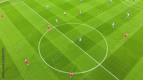Aerial view of football players at the very beginning of the match, the game starting kick-off. On the field, one team is in red, the other is in blue and the referee is in yellow. Taken by drone