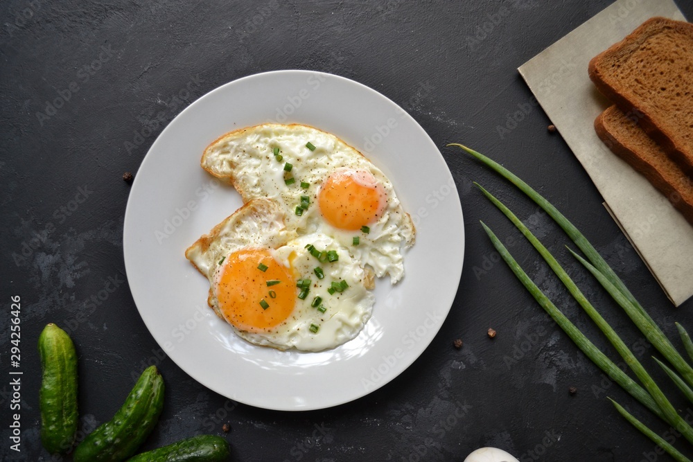 Tasty breakfast. Food on the table. Food on a black concrete decorative background. Fried eggs in a white plate. Eggs, green onions, brown bread, cucumbers.