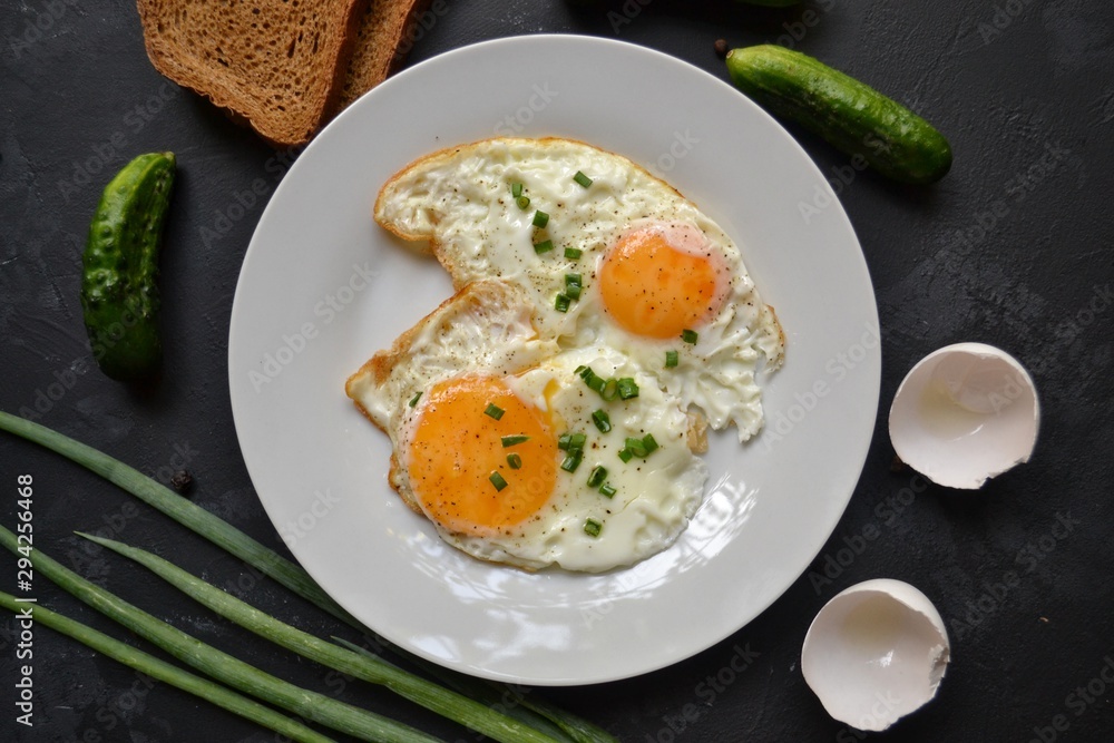 Tasty breakfast. Food on the table. Food on a black concrete decorative background. Fried eggs in a white plate. Eggs, green onions, brown bread, cucumbers.