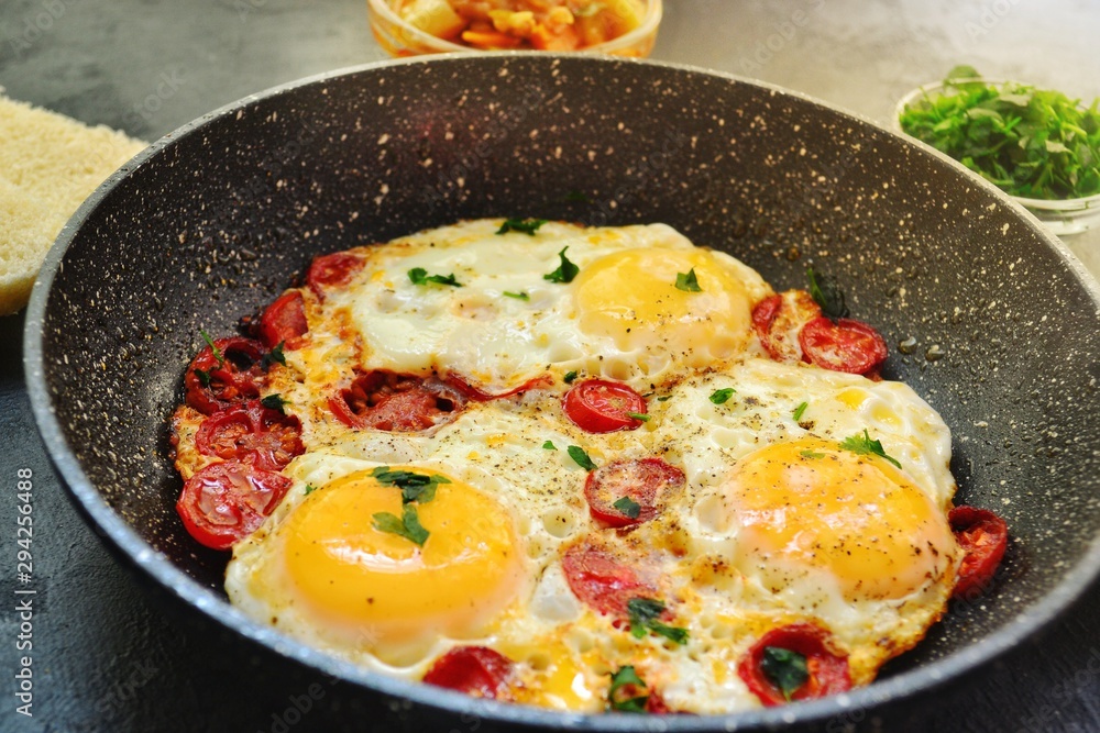 Fried eggs with tomatoes and parsley. Fried eggs in a black pan on a dark background. Scrambled Egg Ingredients Fried eggs with tomatoes and parsley. Fried eggs in a black pan on a dark background. Scrambled Egg Ingredients
