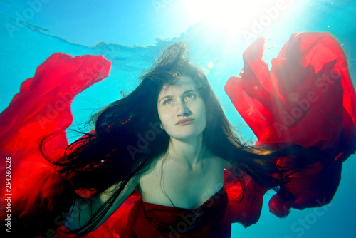 A beautiful, extraordinary girl swims under the water with a red cloth, with her hair down, her arms outstretched like a bird against the background of sunlight from the surface. Portrait. Close up