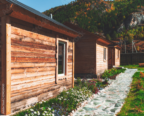 The row of three wooden single-window shacks on the area of a mountain resort or camp with a paved pathway with flowerbeds in front and partly yellowed autumn mountain ridge in a defocused background