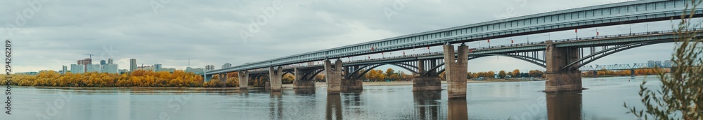 Naklejka premium Panoramic shot of river Ob and its bank in Novosibirsk, Russia during autumn: a metro bridge and transport bridge on the right, the shoreline with yellowed fall trees and cityscape in the background
