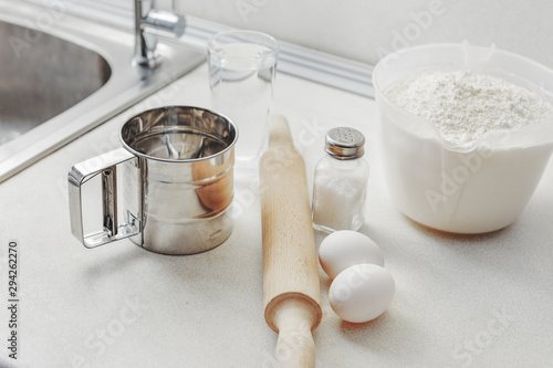 Kitchen table ingredients for making homemade meal from flour. Ingredients for baking fresh bread and cake. Flour, eggs, beater, rolling pin, salt, glass of water and eggshell white table.