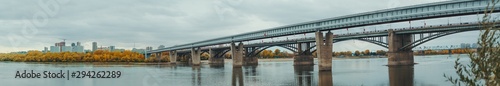 Panoramic shot of river Ob and its bank in Novosibirsk, Russia during autumn: a metro bridge and transport bridge on the right, the shoreline with yellowed fall trees and cityscape in the background © skyNext