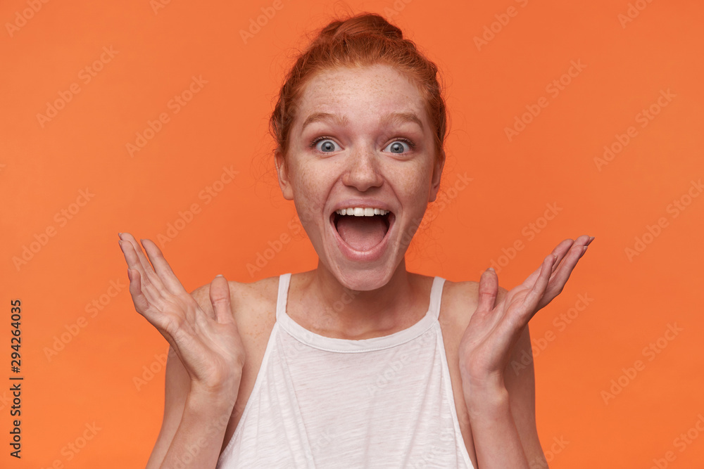 Indoor photo of joyful young readhead female with bun hairstyle wearing white top, looking to camera with surprised face and wide mouth opened, posing over orange background with raised palms Indoor photo of joyful young readhead female with bun hairstyle wearing white top, looking to camera with surprised face and wide mouth opened, posing over orange background with raised palms