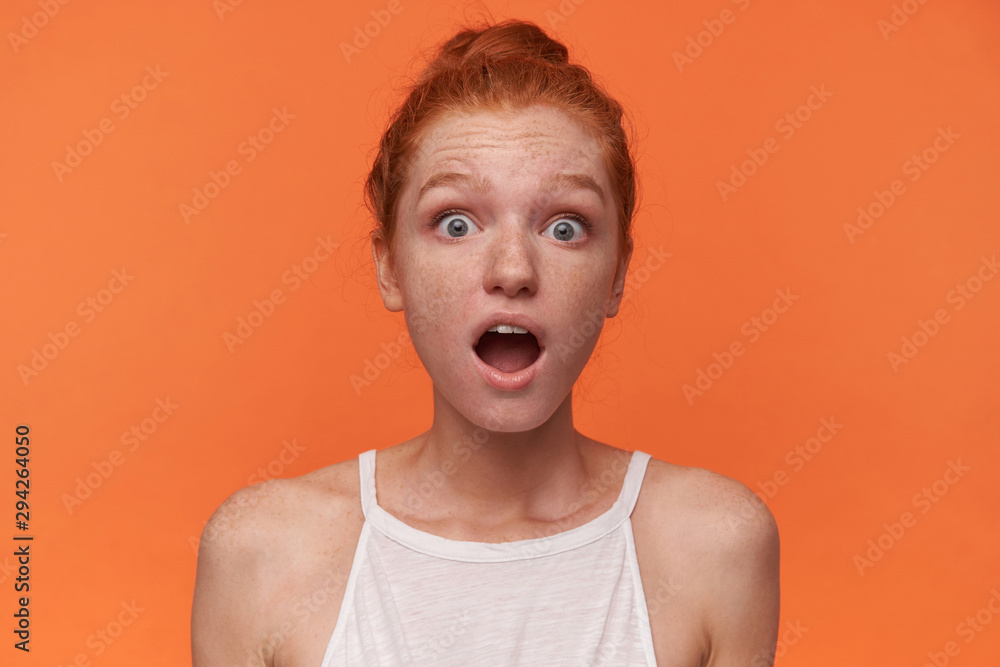 Close-up of surprised young female wearing her red hair in knot over orange background, wearing white top, looking to camera with wide mouth opened and rounding eyes amazedly Close-up of surprised young female wearing her red hair in knot over orange background, wearing white top, looking to camera with wide mouth opened and rounding eyes amazedly