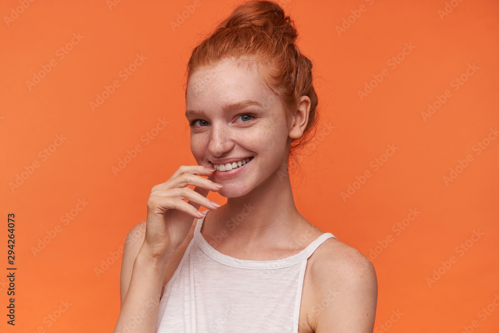 Portrait of positive young redhead woman with knot wearing white top, looking at camera cheerfully and touching underlip with index finger, posing over orange background Portrait of positive young redhead woman with knot wearing white top, looking at camera cheerfully and touching underlip with index finger, posing over orange background