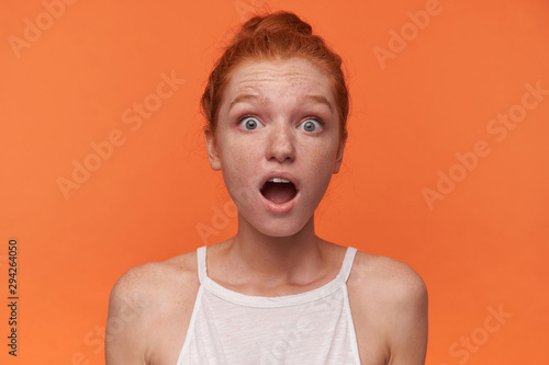 Close-up of surprised young female wearing her red hair in knot over orange background, wearing white top, looking to camera with wide mouth opened and rounding eyes amazedly photo