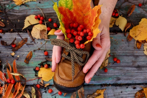 Kid's leather boot with autumn colorful red, orange, yellow maple leaves, ashberries inside  is held by female hands on rustic background. Autumn mood, fall, trendy and stylish concept. photo