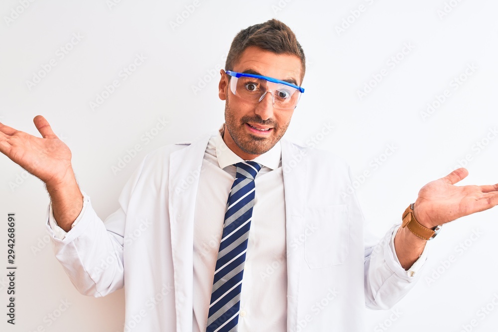 Young handsome scientist man wearing safety glasses over isolated background clueless and confused expression with arms and hands raised. Doubt concept. Young handsome scientist man wearing safety glasses over isolated background clueless and confused expression with arms and hands raised. Doubt concept.