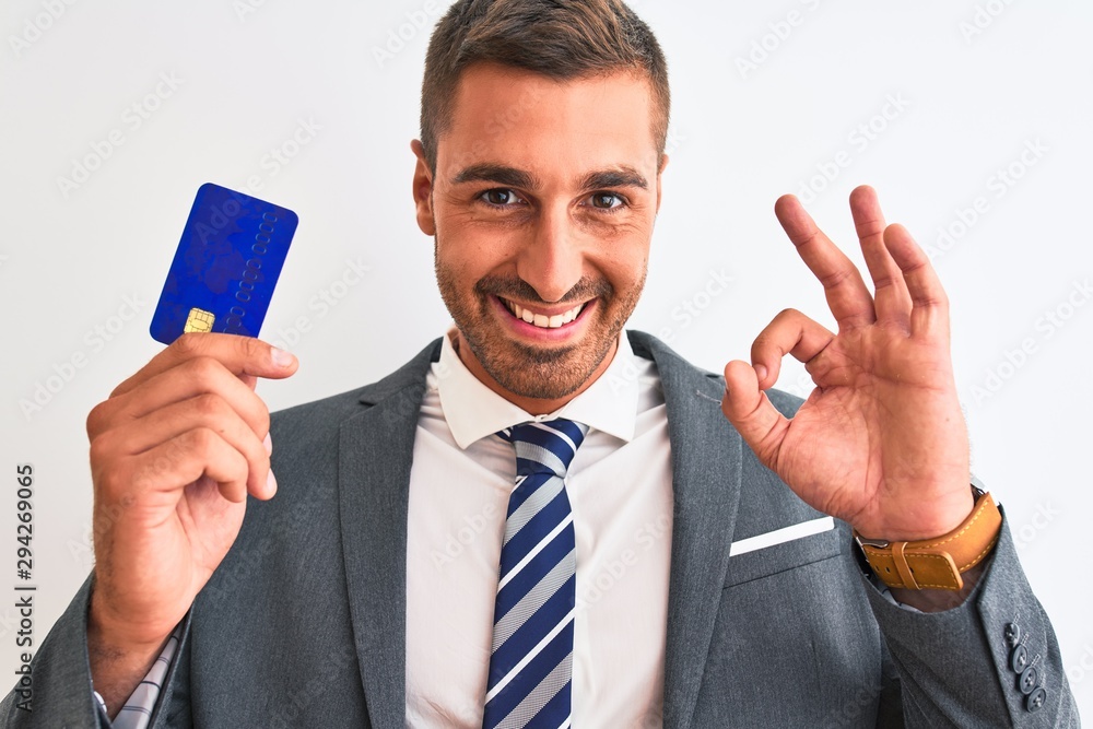 Young handsome business man holding credit card over isolated background doing ok sign with fingers, excellent symbol Young handsome business man holding credit card over isolated background doing ok sign with fingers, excellent symbol