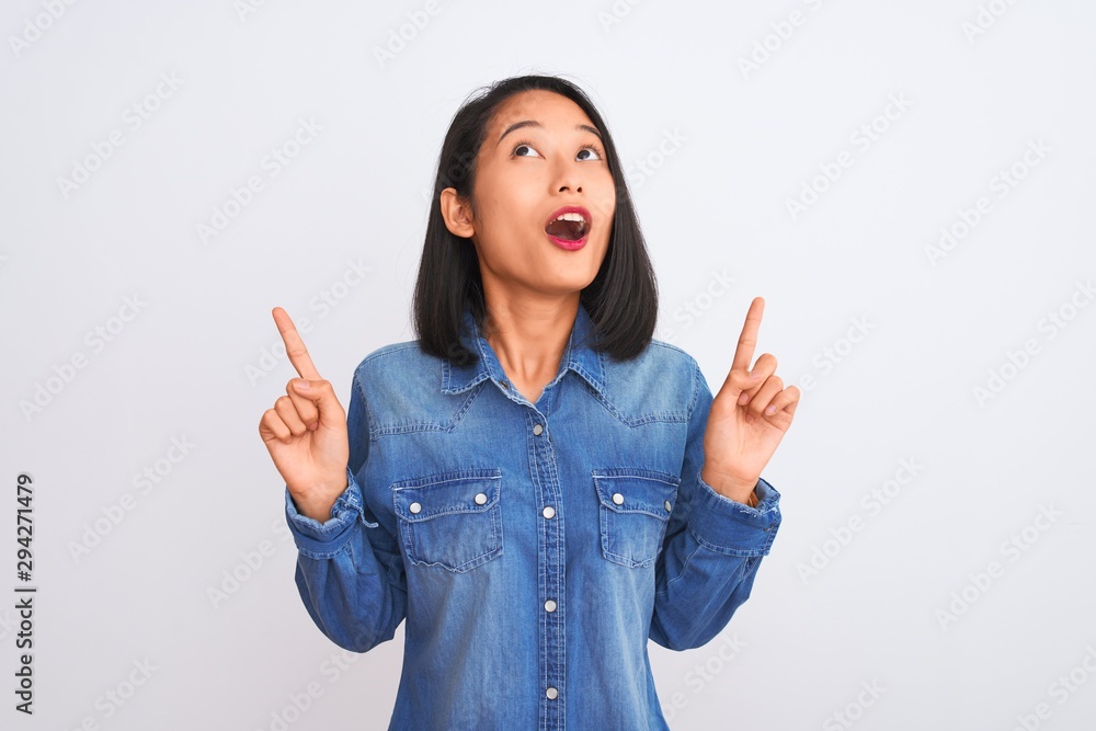 Young beautiful chinese woman wearing denim shirt standing over isolated white background amazed and surprised looking up and pointing with fingers and raised arms. Young beautiful chinese woman wearing denim shirt standing over isolated white background amazed and surprised looking up and pointing with fingers and raised arms.