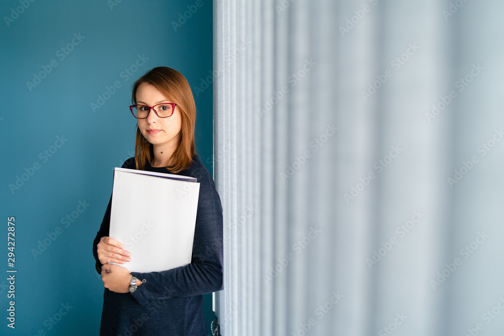 Portrait of young business woman with glasses secretary holding file folder job application in front of blue wall by window curtains Portrait of young business woman with glasses secretary holding file folder job application in front of blue wall by window curtains