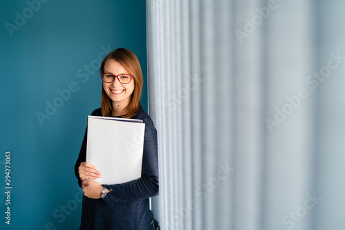 Portrait of young business woman with glasses secretary holding file folder job application in front of blue wall by window curtains smiling
