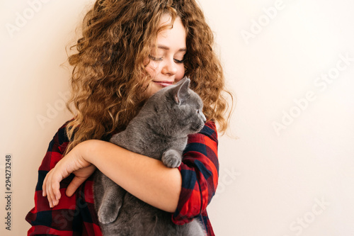 Curly blond teenage girl smiles and holds in her hands a gray kitten of Scottish breed. In a red plaid shirt. Delicate pastel makeup. Model age 12 years