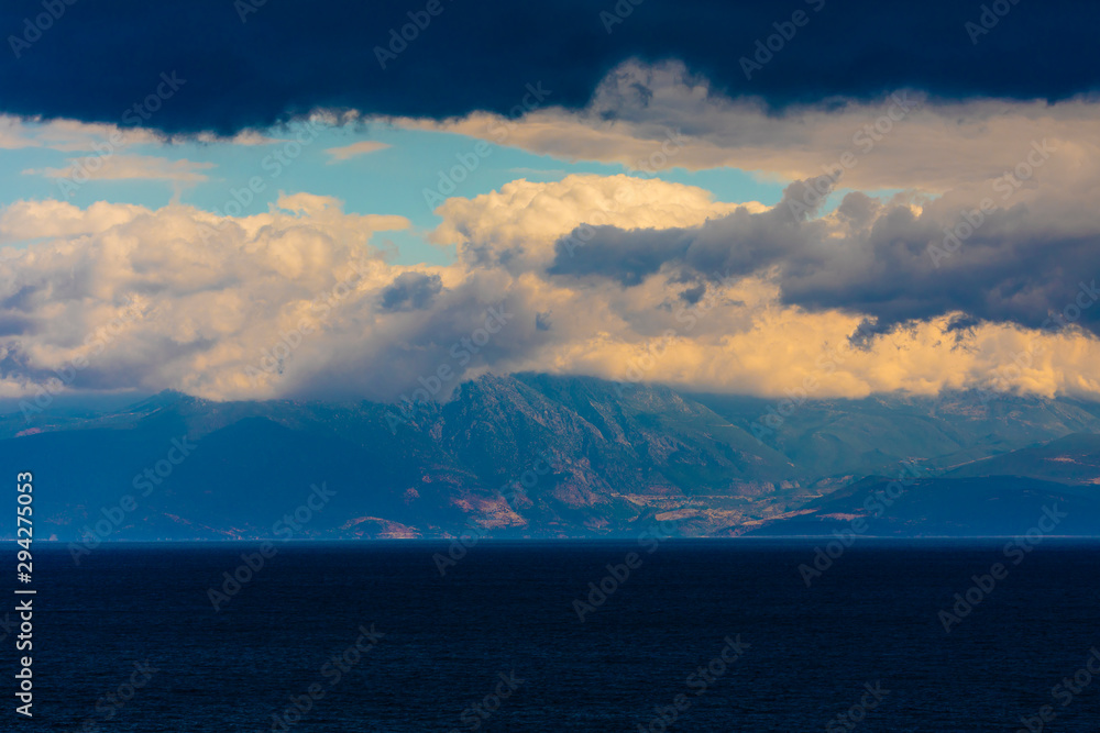 A mountain range in Greece covered in low hanging cloud with the ocean in the foreground.