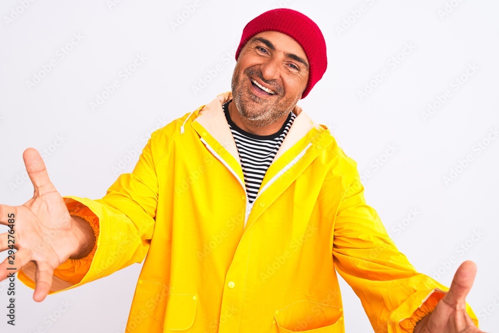 Middle age man wearing rain coat and woolen hat standing over isolated white background looking at the camera smiling with open arms for hug. Cheerful expression embracing happiness.