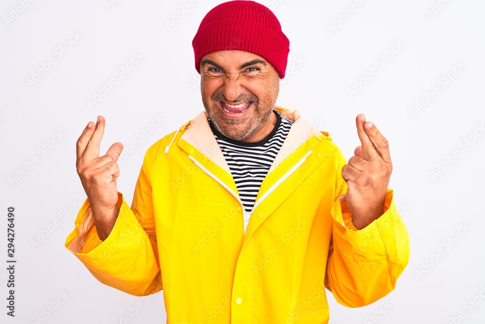 Naklejka premium Middle age man wearing rain coat and woolen hat standing over isolated white background gesturing finger crossed smiling with hope and eyes closed. Luck and superstitious concept.