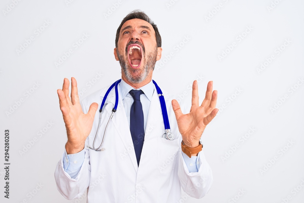 Middle age doctor man wearing coat and stethoscope standing over isolated white background crazy and mad shouting and yelling with aggressive expression and arms raised. Frustration concept. Middle age doctor man wearing coat and stethoscope standing over isolated white background crazy and mad shouting and yelling with aggressive expression and arms raised. Frustration concept.