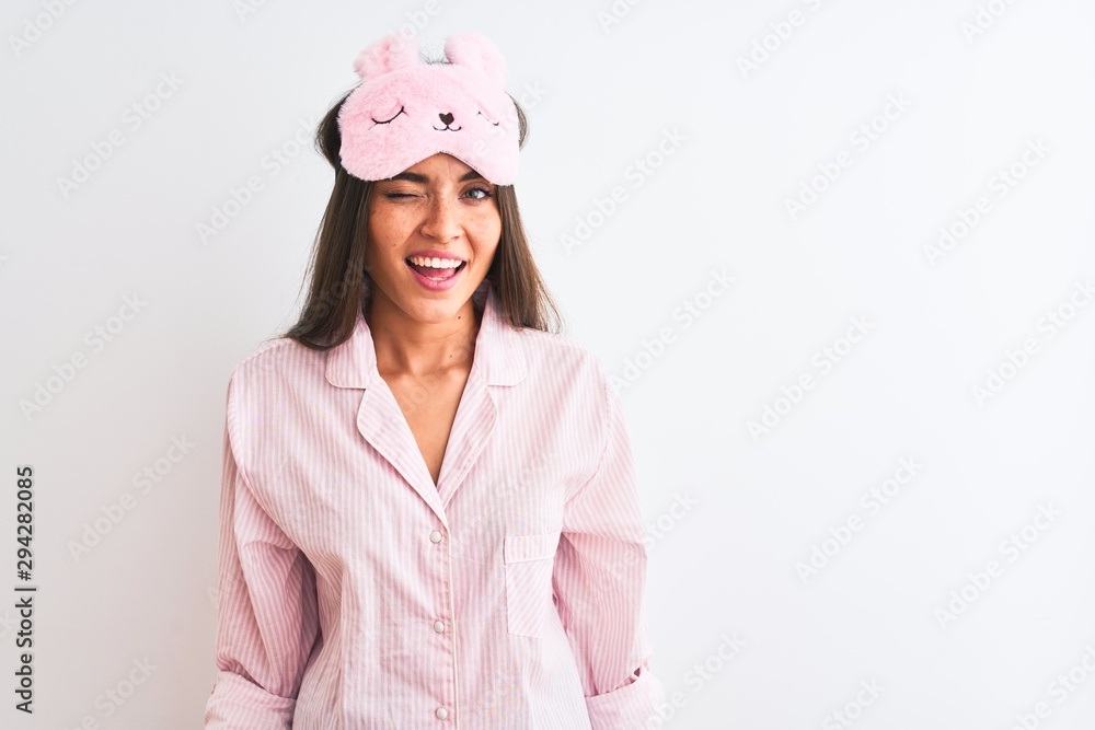 Young beautiful woman wearing sleep mask and pajama over isolated white background winking looking at the camera with sexy expression, cheerful and happy face. Young beautiful woman wearing sleep mask and pajama over isolated white background winking looking at the camera with sexy expression, cheerful and happy face.