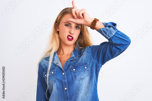 Young beautiful woman wearing casual denim shirt standing over isolated white background making fun of people with fingers on forehead doing loser gesture mocking and insulting.