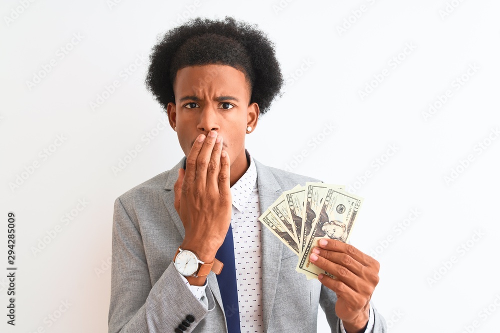 Young african american businessman holding dollars standing over isolated white background cover mouth with hand shocked with shame for mistake, expression of fear, scared in silence, secret concept Young african american businessman holding dollars standing over isolated white background cover mouth with hand shocked with shame for mistake, expression of fear, scared in silence, secret concept