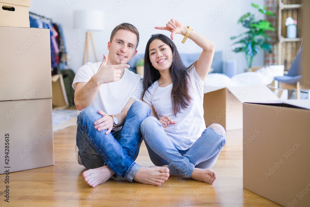 Young beautiful couple sitting on the floor at new home around cardboard boxes smiling making frame with hands and fingers with happy face. Creativity and photography concept. Young beautiful couple sitting on the floor at new home around cardboard boxes smiling making frame with hands and fingers with happy face. Creativity and photography concept.
